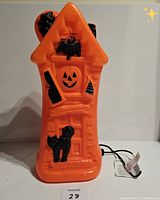 Front view of the orange blow mold haunted house, showing black decorative elements including bats, jack-o'-lantern face, and a black cat, with attached clip light and cord.