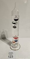 Full view of the vintage Galileo thermometer showing the tall slender glass tube with colorful floating spheres inside and metal tags.