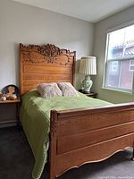 Full view of antique full size bed with green bedspread, wooden carved headboard and footboard, bedside table with lamp in a bedroom.