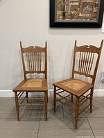 Front angled view of the pair of oak Victorian style spindle back chairs with cane seats against a white wall and tiled floor.