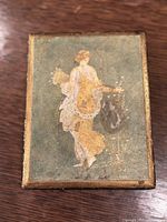 Closed wooden trinket box with an image of a classical female figure, Flora (Khloris), goddess of flowers, on its lid. The image is faded but visible.