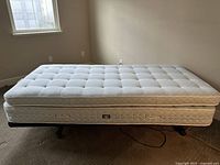 Full view of twin sized mattress on electrical adjustable base with black metal legs in a bedroom corner, mattress tufted on white quilted top.
