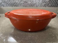 Side profile of closed red cast iron Dutch oven showing shape, lid and handles.