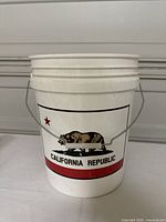 White 5 gallon plastic bucket featuring the California Republic flag design with a bear and star, metal carry handle attached, reinforcing ribs around the bucket.