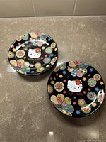 Pair of Hello Kitty ceramic plates displayed on a stone-like surface with colorful floral designs and black background.