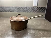 Side view of the small hammered copper pot with lid and metal handle on kitchen counter.