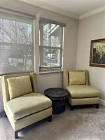 Pair of vintage light green upholstered lounge chairs with wooden legs and decorative pillows, positioned near windows with a small round dark side table between them.