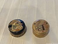 Pair of round handmade jewelry boxes with bird painted lids shown closed on fabric surface.