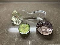 Lot of four vintage paperweights in various shapes on a stone surface: clear whale, geometric prism, floral orb, and spiral design orb.