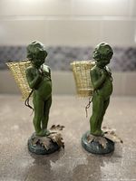 Pair of green cast metal cherub figures with small woven baskets on their backs, standing on bases with leaf designs, photographed from different angles to show details.
