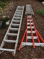 Three ladders laid on ground: an orange ladder, a long aluminum ladder, a shorter aluminum ladder