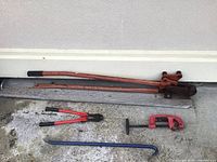 Wide shot showing the four main tools: rebar bender cutter mounted on wood, bolt cutter, pipe cutter, and pry bar arranged on floor against wall.