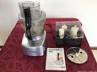 Front view of assembled Cuisinart food processor on a red patterned surface with attachments container and user manual nearby.
