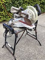 Angle view of Delta Sidekick compound miter saw on metal stand showing saw blade, dust bag with Delta branding, and stand structure.