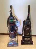 Two upright cleaning appliances standing side-by-side on carpet: Hoover WindTunnel vacuum on right, Hoover SteamVac carpet cleaner on left.