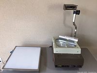 Overview photo showing the Elmo HP-L355 overhead projector with adjustable arm and lens, portable trace light table with illuminated surface, and several rolls of blank transparencies on top of the projector.
