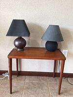 Photo showing a small wooden table with two small lamps on top against a neutral wall and tiled floor. One lamp has a textured dark metal base and another has a smooth black ceramic base, both with dark lampshades.