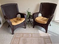 View of two brown high back wicker armchairs with beige cushions, small round black metal table between them with a live cactus on top, and a small beige area rug with a leaf design on floor.