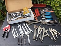 Photo showing assortment of sockets in box, multiple combination wrenches, locking pliers, adjustable wrenches, and other pliers on black surface.
