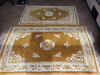 Photo showing two ochre-colored Indian wool area rugs, one larger (76 x 50) and one smaller (38 x 62), laid on a tile floor. Both rugs have a central medallion, corner motifs, floral border, and white fringes.