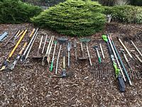 Wide view of all yard tools spread on wood chips in front of bushes. Shows rakes, shovels, post pounder, post digger, and other tools along with the electric hedge trimmer.
