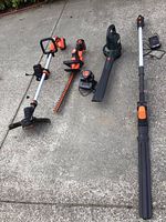 Four yard tools laid out on concrete: Worx trimmer, Black & Decker hedge trimmer, Craftsman blower, Black & Decker weed whacker, with visible batteries and chargers.