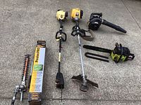 Overall view of all items on concrete: two yellow powerheads, black blower, hedge trimmer and pole saw attachments with box, green chainsaw with bar