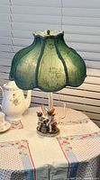 Full view of the antique lamp with green silk shade on a table with floral cloth, showing base and shade.