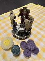 Front and side view of an angel-shaped ceramic candle holder showing four angels holding hands in a circle, with decorative candles placed beside it on a yellow checkered cloth.