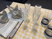 Photo showing 4 clear champagne glasses with vertical etched lines, 2 blue and gold striped vodka cups, 4 beige and blue striped fabric napkins folded in holders, and 4 placemats on yellow checkered tablecloth.