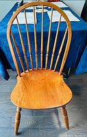 Front view of wooden Windsor chair showing curved top rail, turned spindles, and contoured seat on hardwood floor with a blue cloth surface in the background.