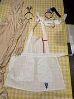 Photo of entire costume set laid flat on yellow checkered surface including beige bodysuit with hood, white apron, and two headbands with accessories.