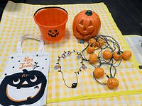 Photo of the complete Halloween decoration set showing all included items laid out on a yellow checkered cloth: pumpkin bucket, ceramic pumpkin lantern, pumpkin LED string lights, skull necklace, and bag.