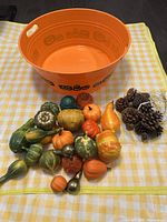 Orange plastic basin with jack-o'-lantern face designs containing artificial pumpkins and pine cones on a yellow checkered cloth.