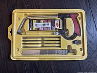 Photo showing the hand jigsaw saw and five different blades in original yellow plastic packaging on dark wood surface.