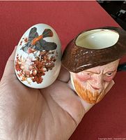 Two ceramic collectibles held in a hand: one a Lancaster Toby mug with a detailed face and hat; the other an Avon decorative egg showing bird and floral designs.