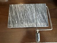 Rectangular grey marble cheese board with white veining placed on wooden surface. Attached metal wire slicer with matching marble handle shown horizontally across the board.