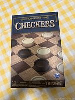 Front view of Traditions Checkers board game box showing checkered board and pieces in black and cream.