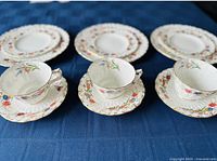 3 teacups and 3 dinner plates with floral and gold trim arranged in a row on a blue cloth