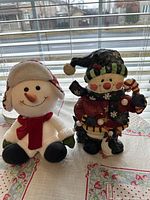 Both snowman decorations placed side by side on a floral patterned cloth backdrop with window blinds behind