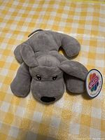 Front view of gray plush dog lying on checkered yellow and white surface showing face and tag
