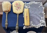Hairbrush, hand mirror, small case, and lace cloth arranged on a blue surface showing needlepoint floral designs and ornate metal handles.