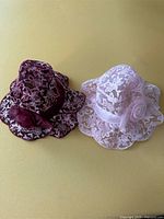 Photo shows two vintage-style lace hats side by side on a neutral background. One hat is maroon with white floral lace overlay and a maroon rose accessory. The other is pale pink with white floral lace overlay and a pink rose on a satin band.