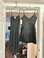 Two black cocktail dresses hanging on hangers on a door, showing one long floral patterned satin-like dress and one short textured polka dot dress.
