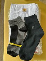 Photo showing gray and black socks with yellow stripe and a plain black sock on white folded t-shirt in plastic packaging.