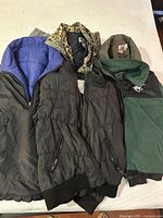 Five jackets displayed on a bed. Three jackets visible in front, including two black puffer vests and one blue quilted jacket, plus partially visible animal print jacket and green fleece-lined jacket.