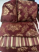 Photo of folded comforter, pillows, and shams showing burgundy and gold damask floral pattern jacquard fabric and striped detail on one item.