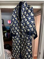 Front view of a blue quilted robe with a cream and yellow floral pattern, hanging from a hanger showing full garment length and sleeve detail.