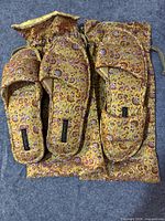 Two small silk slippers with floral embroidery placed on top of a matching silk drawstring pouch, all in gold and purple floral design.