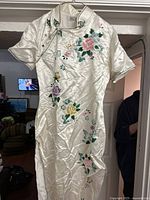White traditional Chinese silk dress hanging on a hanger against a doorway, showing floral embroidery on front and design details.